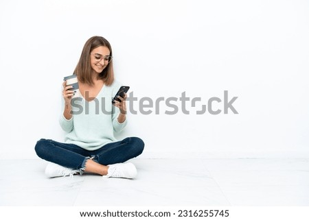 Young caucasian woman sitting on the floor isolated on white background holding coffee to take away and a mobile