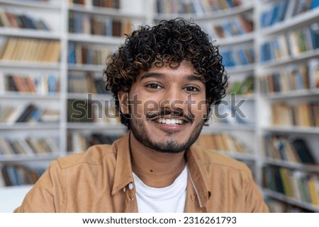 Close-up photo. Face portrait of a young hispanic male student sitting and studying in the library looking at the camera with a smile, chatting online on a video call, recording a podcast.