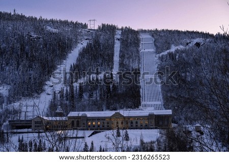 A scenic winter view of the Rjukan Norway hydroelectric power station, its surrounding snow-covered mountains, and the nearby frozen lake