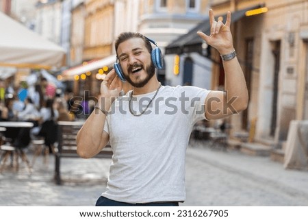 Happy relaxed overjoyed bearded young man in wireless headphones choosing, listening favorite energetic disco rock n roll music dancing outdoors. Guy walking in urban sunshine crowded city street Royalty-Free Stock Photo #2316267905
