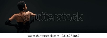 Side view of trendy african america model in elegant gloves and golden accessories touching hips while standing isolated on black, high fashion and evening look, banner
