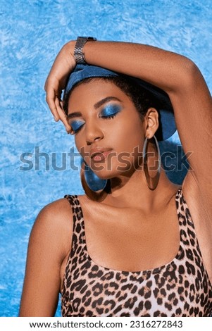 Portrait of trendy african american woman with bold makeup and golden earrings posing in top with animal print and touching beret on blue textured background, stylish denim attire