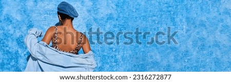 Back view of young african american woman posing in beret, top with animal print and denim shirt while standing and posing on blue textured background, stylish denim attire, banner