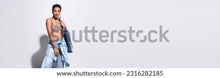 Trendy young african american model with short hair posing in top with animal print and denim clothes on grey background with copy space, denim fashion concept, banner Royalty-Free Stock Photo #2316282185