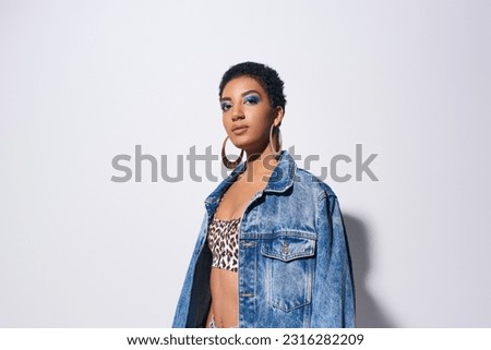 Portrait of confident african american model with bold makeup and golden earrings looking at camera while posing in top with animal print and denim jacket on grey background, denim fashion concept