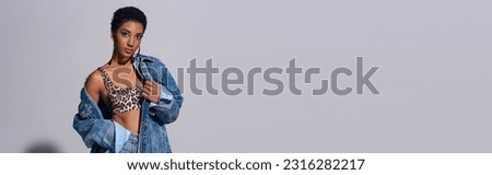 Modern short haired african american woman in top with animal print and denim clothes looking at camera and standing on grey background, denim fashion concept, banner Royalty-Free Stock Photo #2316282217
