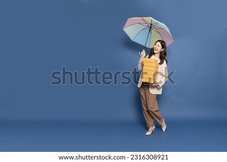 Happy Young Asian woman standing and holding package parcel box and rainbow umbrella isolated on blue background, Delivery courier and shipping service in rainy season concept