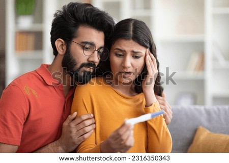 Unwanted Pregnancy. Worried young indian couple looking at positive test result, stressed millennial eastern man and woman sitting on couch at home, not ready to be parents, copy space