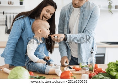 Close up of cute infant girl savouring juicy vegetable given by loving parent from cutting board in modern kitchen. Cheerful caucasian family benefiting from healthy dinner in home interior. Royalty-Free Stock Photo #2316350553
