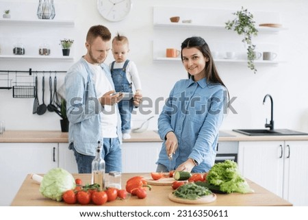 Charming brunette woman posing at kitchen table while calm father holding cell phone and baby girl at worktop. Young parents searching for dinner ideas with fresh vegetables using internet connection.
