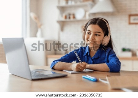 Digital Education. Happy Schoolgirl At Laptop Engages In Online Learning And Active Note Taking Writing In Textbook, Doing Homework At Home, Wearing Headphones. E-Learning Routine Concept