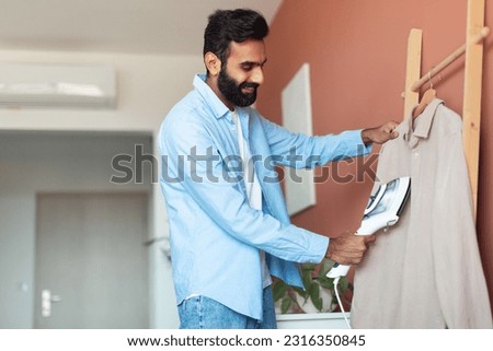 Happy Arabic Man Using Clothes Steamer Ironing And Steaming His Shirt Standing At Modern Home Interior. Guy Caring For Clothing. Male Wardrobe Maintenance And Domestic Devices For Comfort