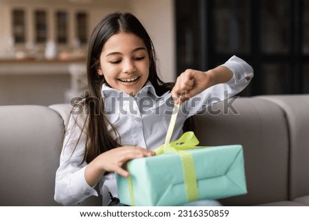 Birthday Gift. Happy Middle Eastern Kid Girl Opening Present Box Celebrating Holiday Sitting On Sofa At Home. Child Posing With Wrapped Gift. B-Day Celebration Concept. Selective Focus