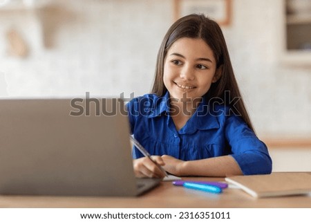 Online Homework. Smiling Hispanic Schoolgirl Writing Notes In Workbook Looking At Laptop Computer, Studying Online And Having Virtual Class At Home, Sitting At Table Indoors. E-Learning Concept