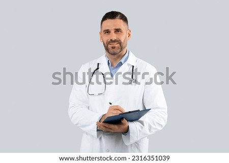 Professional doctor man in medical coat with clipboard in hands posing over light grey wall background, middle aged physician with stethoscope looking at camera Royalty-Free Stock Photo #2316351039