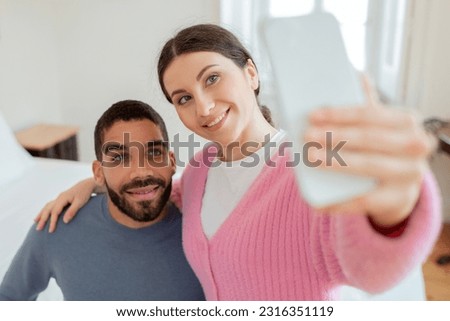 Social Media Presence. Cheerful Bloggers Couple Making Selfie Holding Smartphone In Hand, Posing Embracing In Modern Bedroom Interior. Selective Focus On Young Spouses. Gadgets And Online Fun