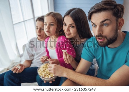 Beautiful young parents, their  daughter and son are watching TV, eating popcorn and showing surprise, sitting on sofa at home Royalty-Free Stock Photo #2316357249