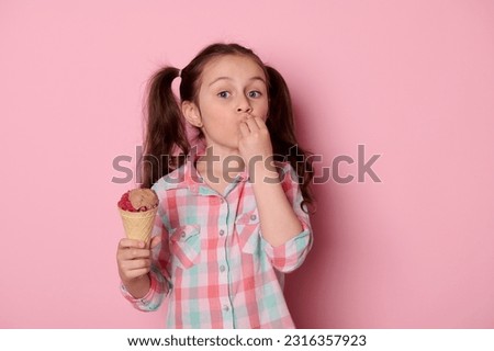 Emotional beautiful little child girl with two ponytails, holding a waffle cone with ice cream, kissing finger and showing delicious hand sign looking at camera, isolated over pink color background