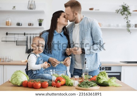 Adorable baby on kitchen table holding red vegetable while loving couple kissing in kitchen interior. Affectionate parents and daughter creating emotional family ties while cooking together at home.