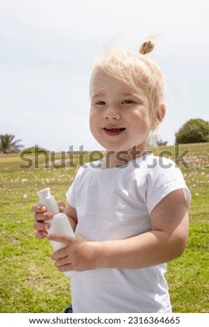 Cute blond child holding  sunscreen while standing in the park, green grass on the background. Sunny day. Smiling face, positive boy 2 years old. Concept of skin protection, summer, parenthood.