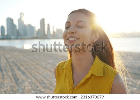 Close-up of young woman on sand beach relaxing breathing fresh air at sunset with city skyscrapers on the background