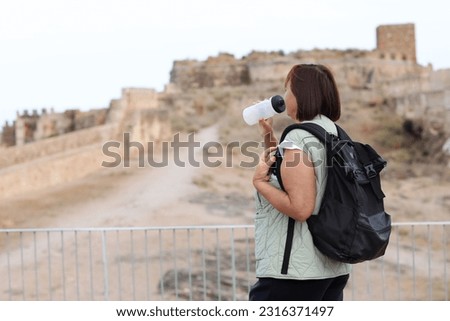 Adult traveling woman tourist with backpack drinks water while relax after walks, posing outdoors in ancient Europe fortress ruins. Retired people summer holiday vacation, active lifestyle concept. Royalty-Free Stock Photo #2316371497