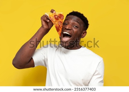 happy african american man in white t-shirt holds slice of pizza and opens his mouth on yellow isolated background, young guy eats fast food and puts pizza in his mouth Royalty-Free Stock Photo #2316377403
