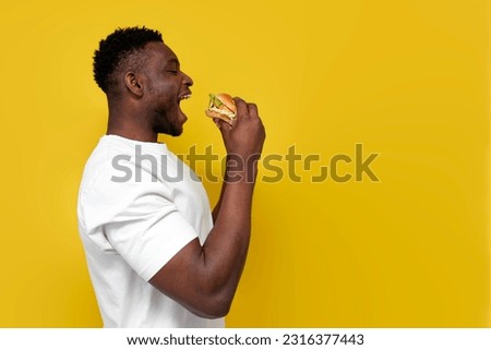 african american man in white t-shirt holds big burger with his mouth open, the guy eats fast food from the side on yellow isolated background, copy space Royalty-Free Stock Photo #2316377443