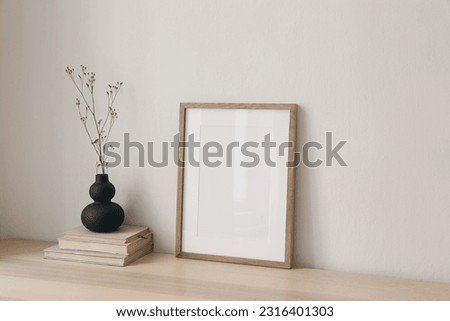 Black organic shaped vase with dry flowers, grass on old books. Blank picture frame mockup, wooden table at home. Minimal Scandi boho interior. Beige wall background. Empty copy space. Side view.