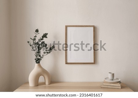 Empty vertical picture frame mockup hanging on beige wall. Cup of coffee, books on wooden table. Vase with silver eucalyptus tree branches. Minimal working space, home office. Scandinavian interior.