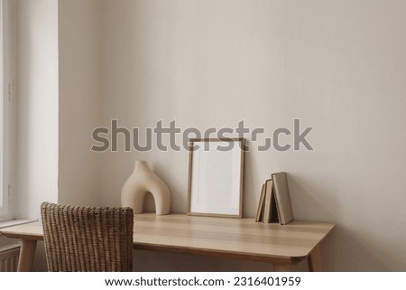Blank picture frame mockup on wooden table, desk with rattan chair. Modern organic shaped vase and old books. Minimal Scandi boho interior. Beige wall background. Elegant minimal interior, home decor.