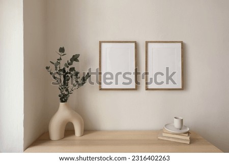 Two vertical picture frame mockup hanging on beige wall. Cup of coffee, books on wooden table. Vase with silver eucalyptus tree branches. Elegant interior, beautiful home decor. Trendy working space.