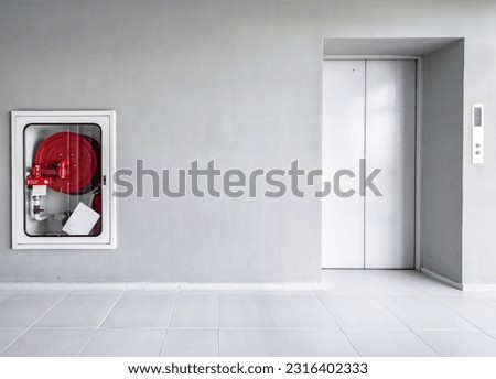 Red fire hose reel in glass cabinet wall at the corridor next to elevator in condominium, building laws and regulations safety requirement inside the area. Fire extinguisher near the closed lift. Royalty-Free Stock Photo #2316402333