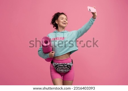smiling happy beautiful woman in stylish sport outfit posing on pink background isolated in studio, wearing blue hoodie, bag and shorts sportswear, holding yoga mat, using smartphone, taking selfie Royalty-Free Stock Photo #2316403393