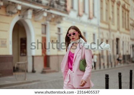 Fashionable elegant confident woman wearing trendy pink sunglasses, suit blazer, white silk scarf, trousers, with green shoulder bag, walking in street of European city. Copy, empty space for text Royalty-Free Stock Photo #2316405537