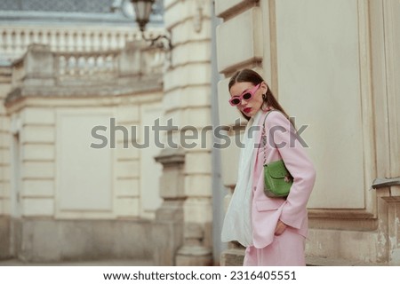 Fashionable elegant confident woman wearing trendy pink sunglasses, suit blazer, white silk scarf, trousers, with green faux leather bag, posing in street of European city. Outdoor fashion portrait Royalty-Free Stock Photo #2316405551