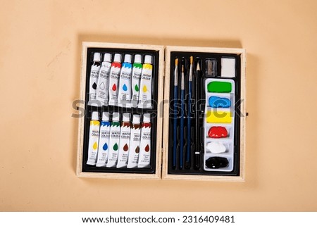 Landscape image of a birds eye view of a watercolour paint set in a wooden box on a beige background. There is three paintbrushes, one led pencil, a sharpener, an eraser and paint pallette