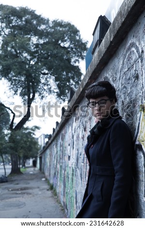 A retro styled girl leaning against a graffiti sprayed wall in a urban environment.