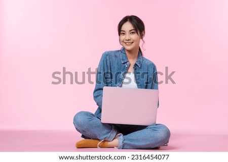 Young Asian college student sits cross-legged on floor working with a laptop computer, looking directly at the camera in a studio shot on a pink background