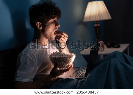 Young guy sits on the bed at night, enthusiastically watches TV series on laptop and eating popcorn. Portrait. Copy space Royalty-Free Stock Photo #2316488437