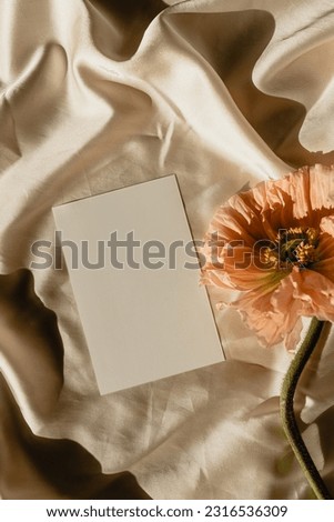 Blank paper sheet card with mockup copy space, poppy flower with aesthetic sunlight shadows on crumpled golden fabric. Aesthetic wedding invitation template. Flat lay, top view
