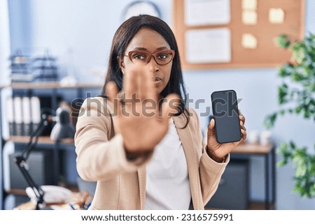 African young woman holding smartphone showing blank screen with open hand doing stop sign with serious and confident expression, defense gesture 