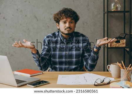 Clueless sad confused mistaken employee business Indian man he wears casual blue checkered shirt shrugging shoulders looking puzzled look camera sit work at office desk with laptop pc computer indoors Royalty-Free Stock Photo #2316580361