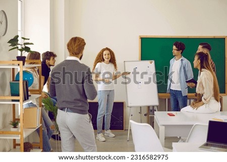 Student girl presenting her project in class. Group of school, college or university students together with teacher listening to young woman giving presentation in front of white board in classroom