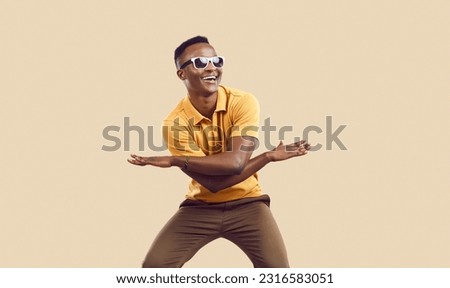 Positive cool dark-skinned man rejoices, has fun and dances isolated on background of mocha color. Funny african american man in casual clothes and sunglasses dancing waving his arms and laughing. Royalty-Free Stock Photo #2316583051