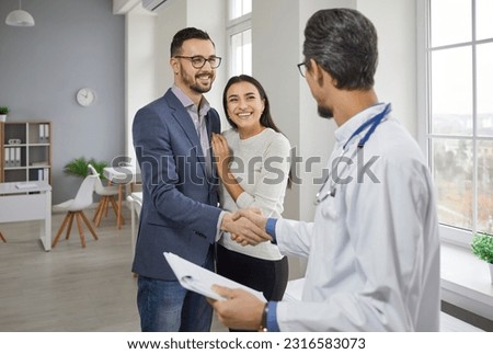 Happy couple meeting family doctor medical specialist in office. Clinician handshaking male patient hand at meeting. Male doctor talking to young husband and wife at health center or hospital Royalty-Free Stock Photo #2316583073