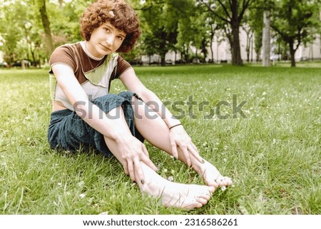 Portrait attractive happy smiling teenager girl, with curly brown hair, brown eyes, sitting on grass in park, barefoot, wearing shorts and T-shirt, summer season, sunny weather