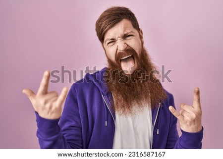 Caucasian man with long beard standing over pink background shouting with crazy expression doing rock symbol with hands up. music star. heavy concept. 