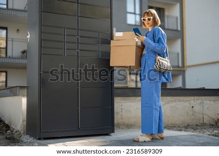 Young woman picks up parcels from automatic post office machine, standing with phone near apartment building. Concept of fast delivery to automatic self lockers Royalty-Free Stock Photo #2316589309
