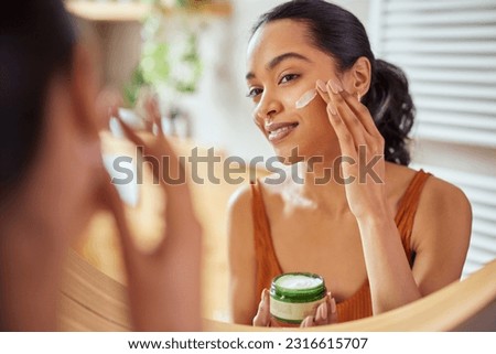 Woman caring of her beautiful skin on the face standing near mirror in the bathroom. Mexican woman applying moisturizer on her face at home. Multiethnic girl holding little green jar of bio skin cream Royalty-Free Stock Photo #2316615707